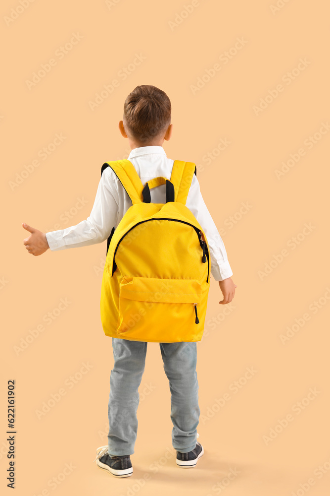 Little schoolboy with yellow backpack showing thumb-up gesture on orange background, back view