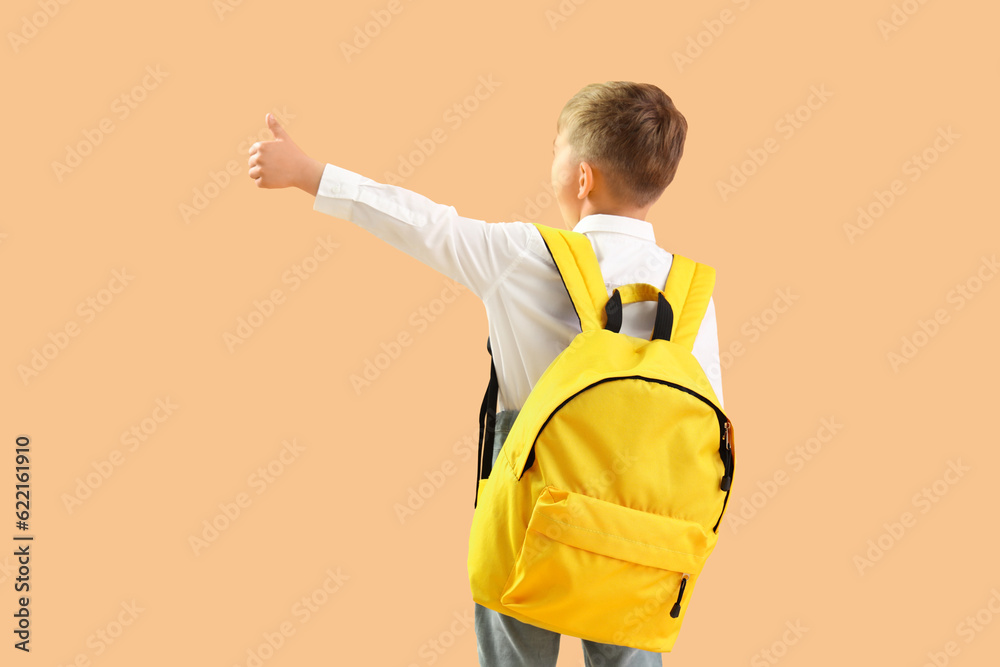 Little schoolboy with yellow backpack showing thumb-up gesture on orange background, back view