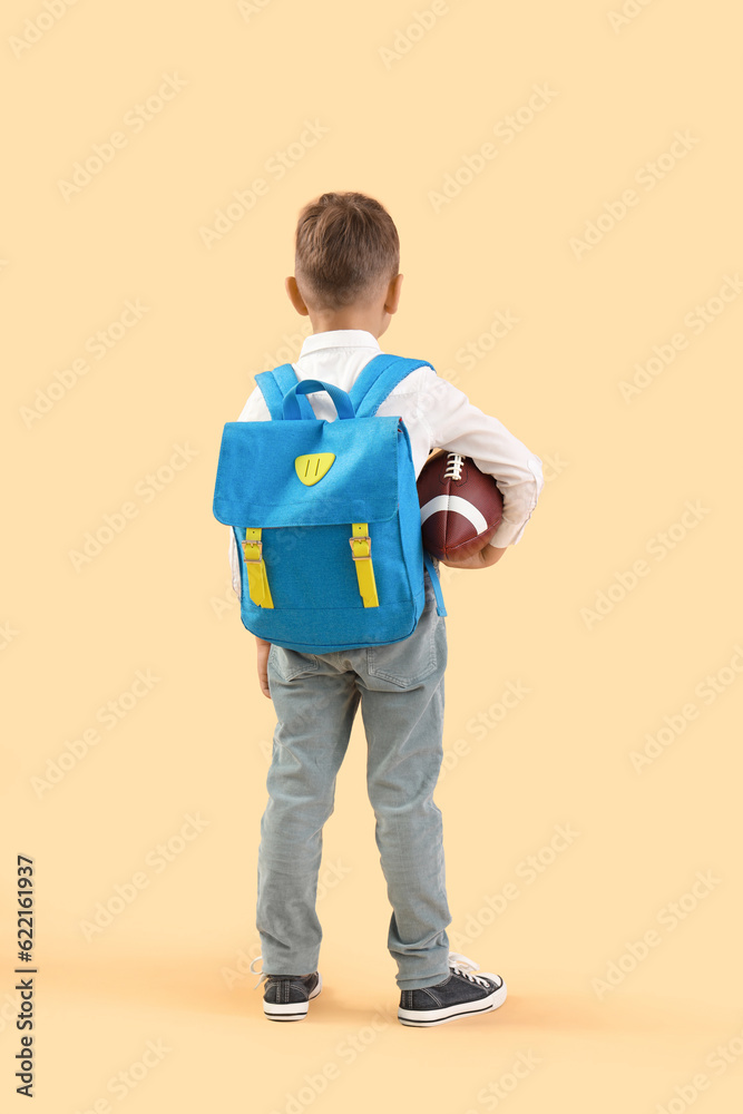 Little schoolboy with blue backpack and rugby ball on orange background, back view