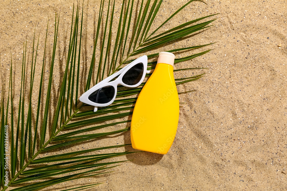 Sunglasses with bottle of sunscreen cream and palm leaf on sand