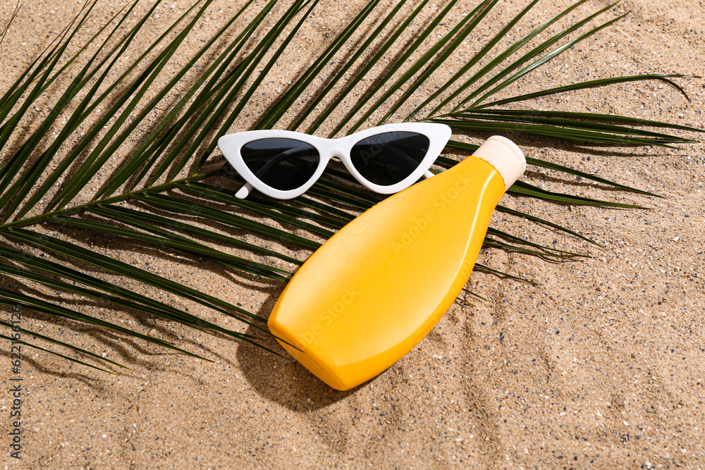 Sunglasses with bottle of sunscreen cream and palm leaf on sand