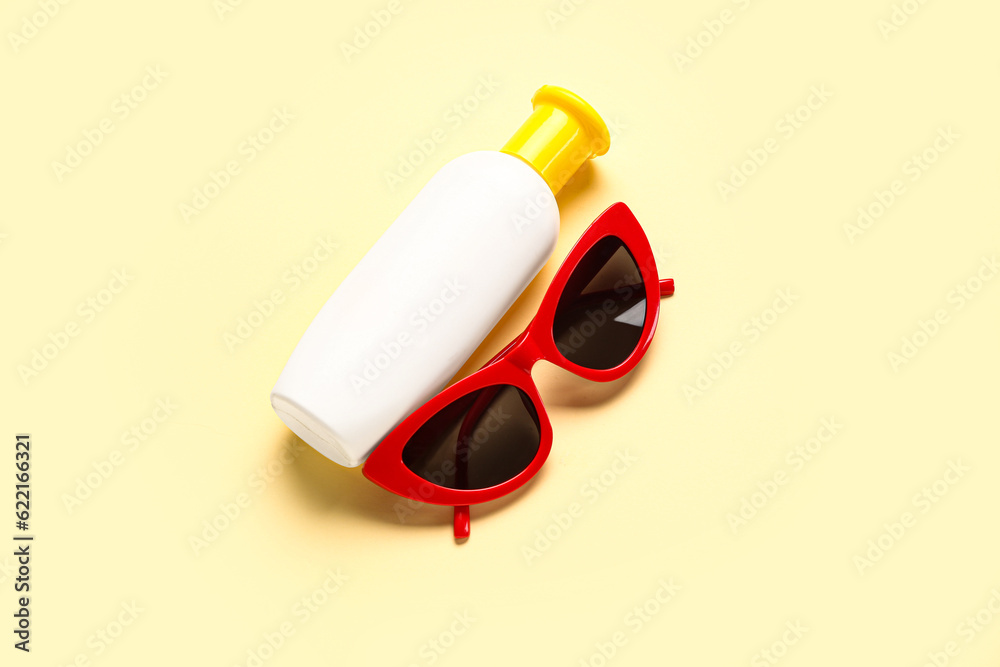 Sunglasses with bottle of sunscreen cream on pale yellow background