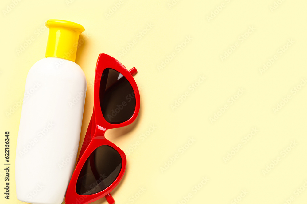 Sunglasses with bottle of sunscreen cream on pale yellow background