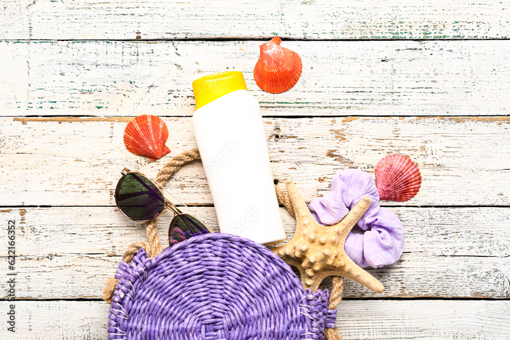 Wicker bag with sunglasses, hair band and bottle of sunscreen cream on white wooden background