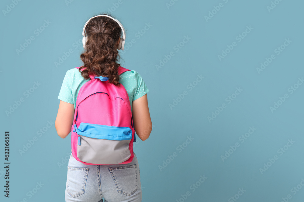 Female student in headphones with backpack on blue background, back view