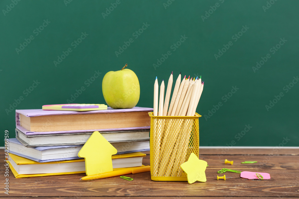 Different stationery with fresh apple on wooden table against green chalkboard