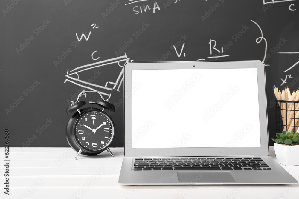 Stationery, alarm clock and laptop on white table against black chalkboard with formulas