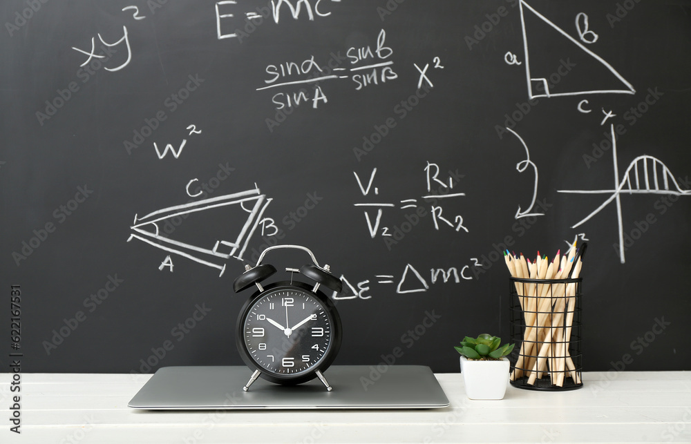 Stationery, alarm clock and laptop on white table against black chalkboard with formulas