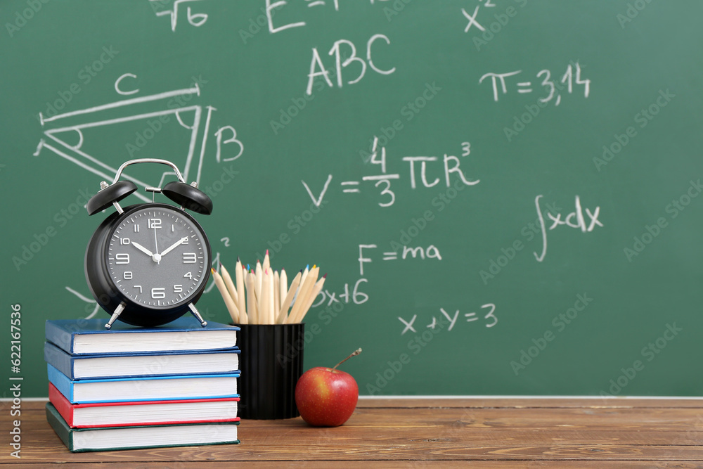 Stationery, alarm clock and fresh apple on wooden table against green chalkboard with formulas