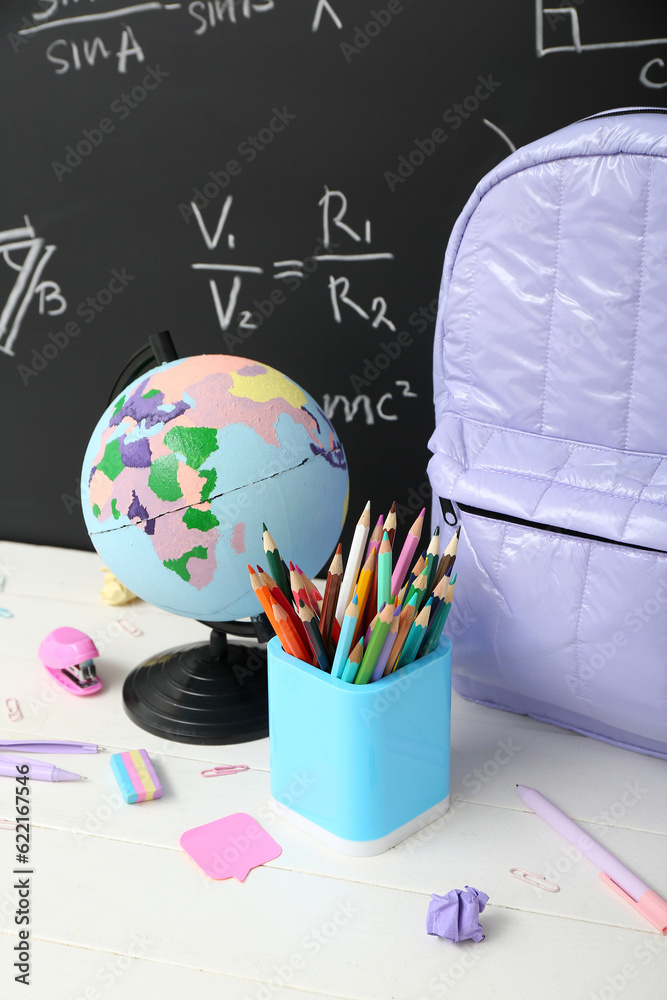 Backpack, stationery and globe on white table against green chalkboard with formulas