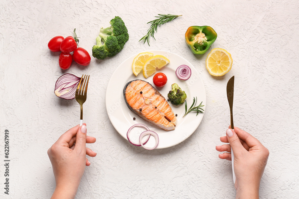 Woman eating tasty grilled salmon steak with vegetables on light background