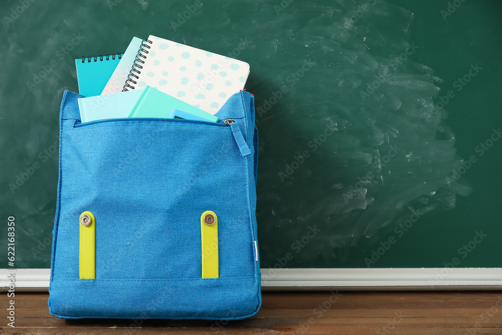 Blue school backpack with notebooks on wooden table near green chalkboard