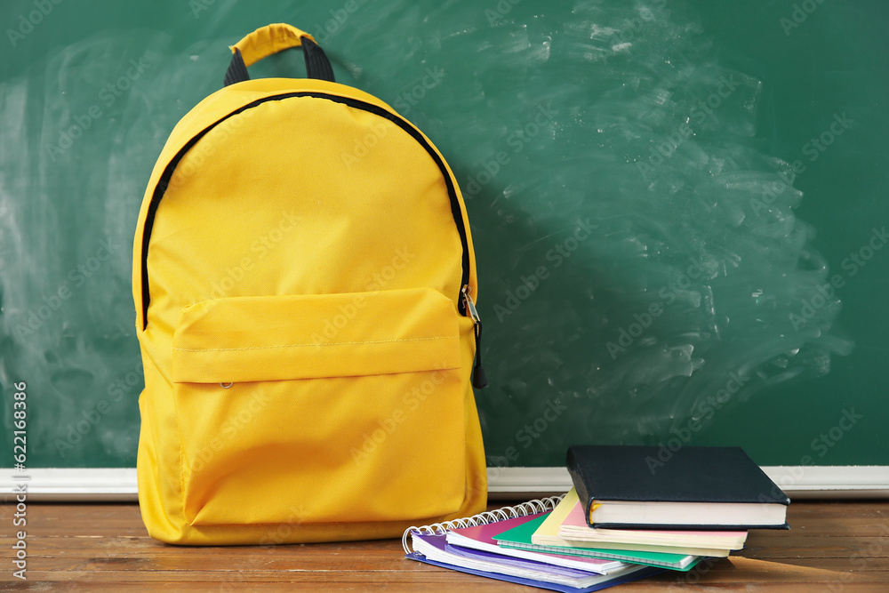 Yellow school backpack with notebooks on wooden table near green chalkboard