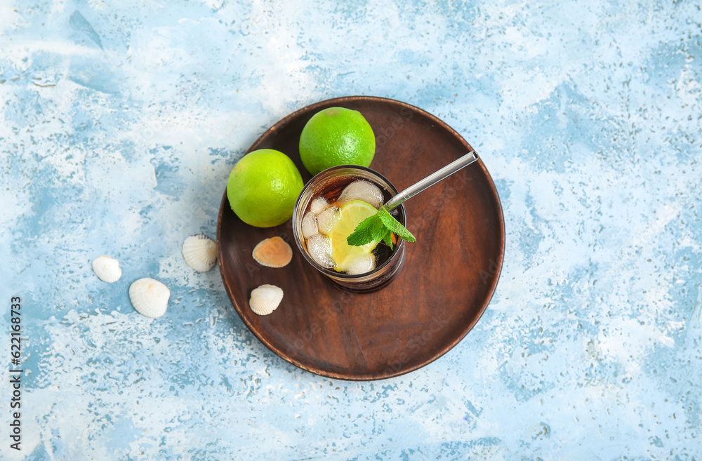 Plate with glass of cold Cuba Libre cocktail and seashells on blue background