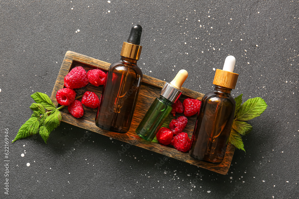 Wooden tray with bottles of cosmetic raspberry oil on black background