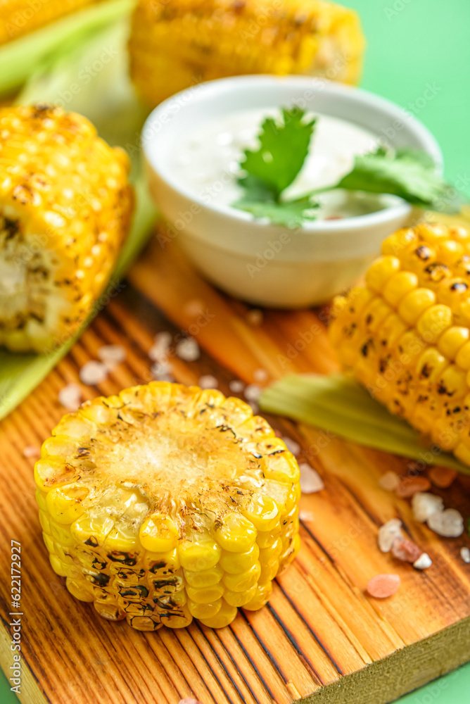 Wooden board with tasty grilled corn and sauce cobs on green background