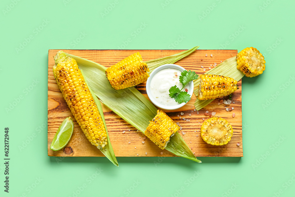 Wooden board with tasty grilled corn and sauce cobs on green background