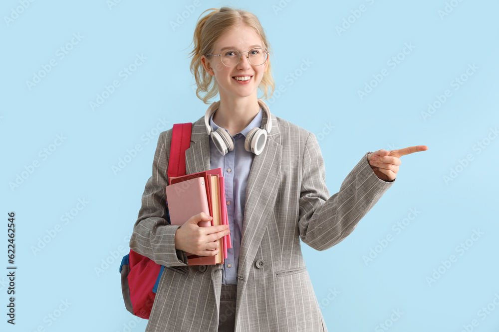 Happy female student with backpack, books and headphones pointing at something on blue background