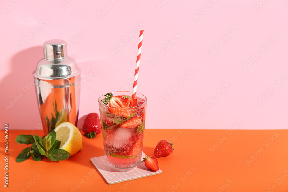 Glass of fresh strawberry mojito with straw and shaker on orange table near pink wall