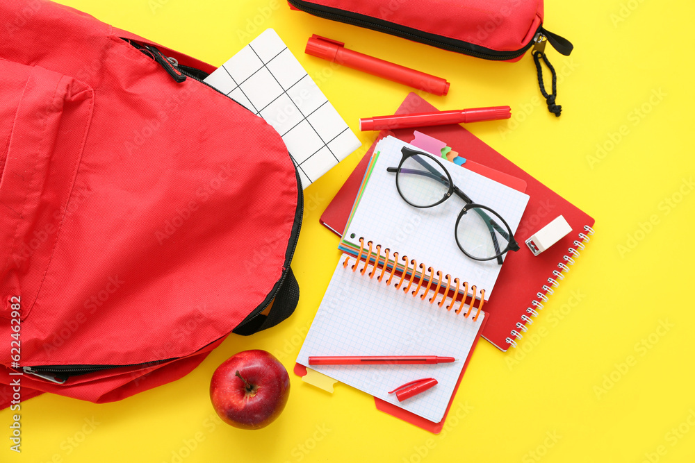 Red school backpack with notebooks, eyeglasses and pencil case on yellow background