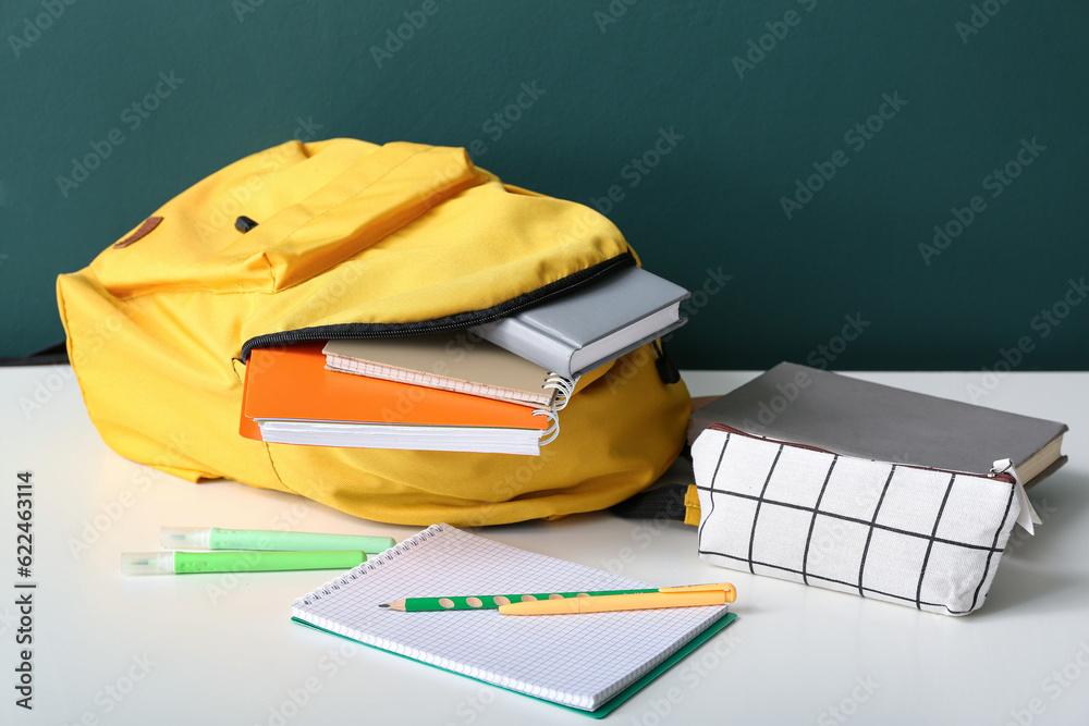 Yellow school backpack with notebooks, pencil case and markers on white wooden table near dark green