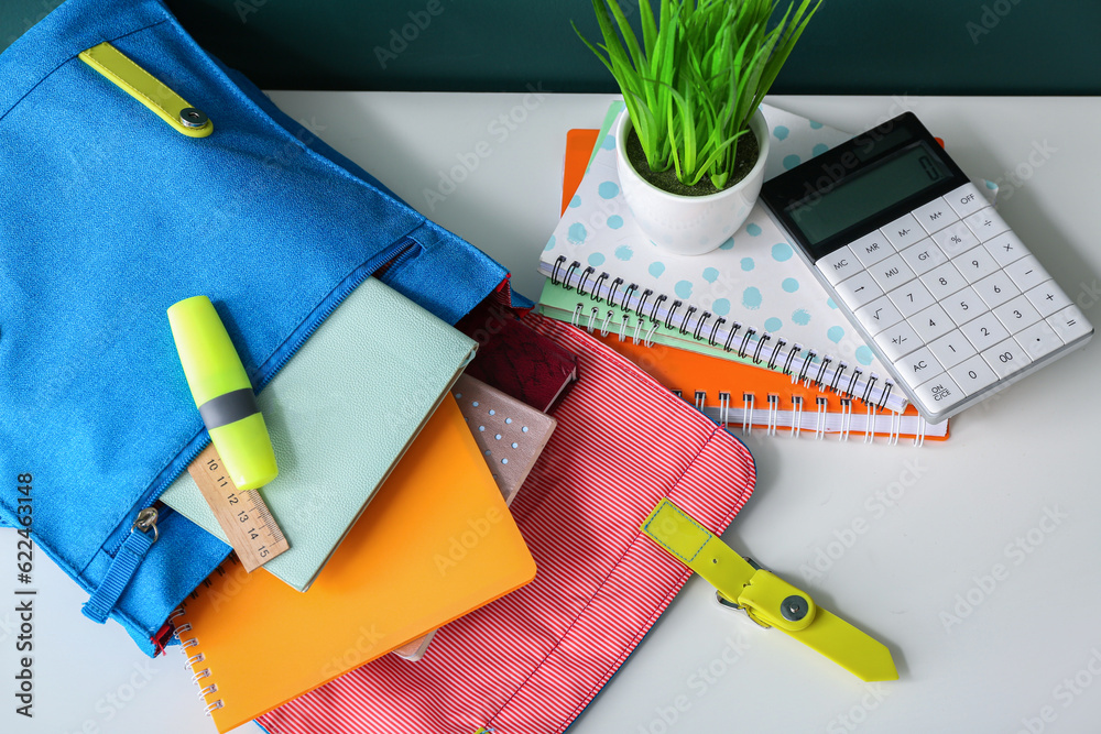 Blue school backpack with notebooks, calculator and houseplant on white wooden table near dark green