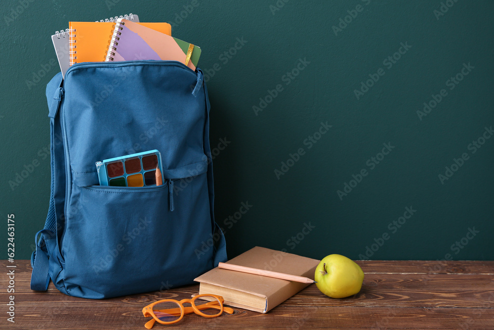 Blue school backpack with notebooks, eyeglasses and apple on wooden table near dark wall