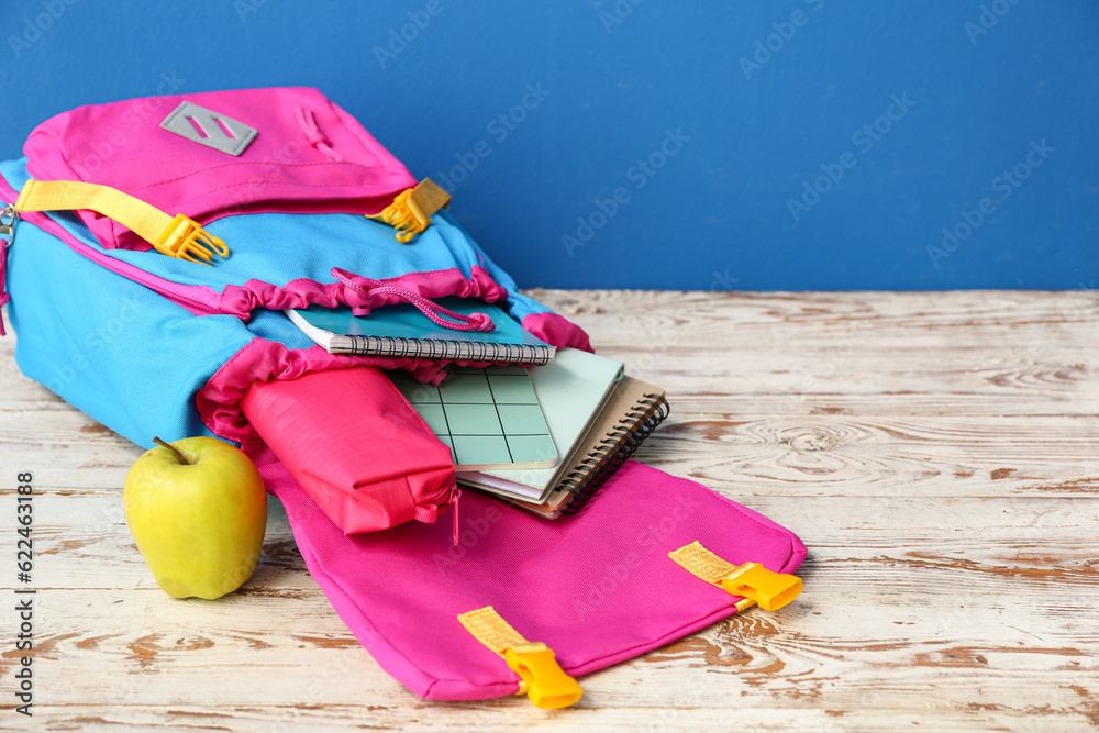 Color school backpack with notebooks, pencil case and apple on wooden table near blue wall