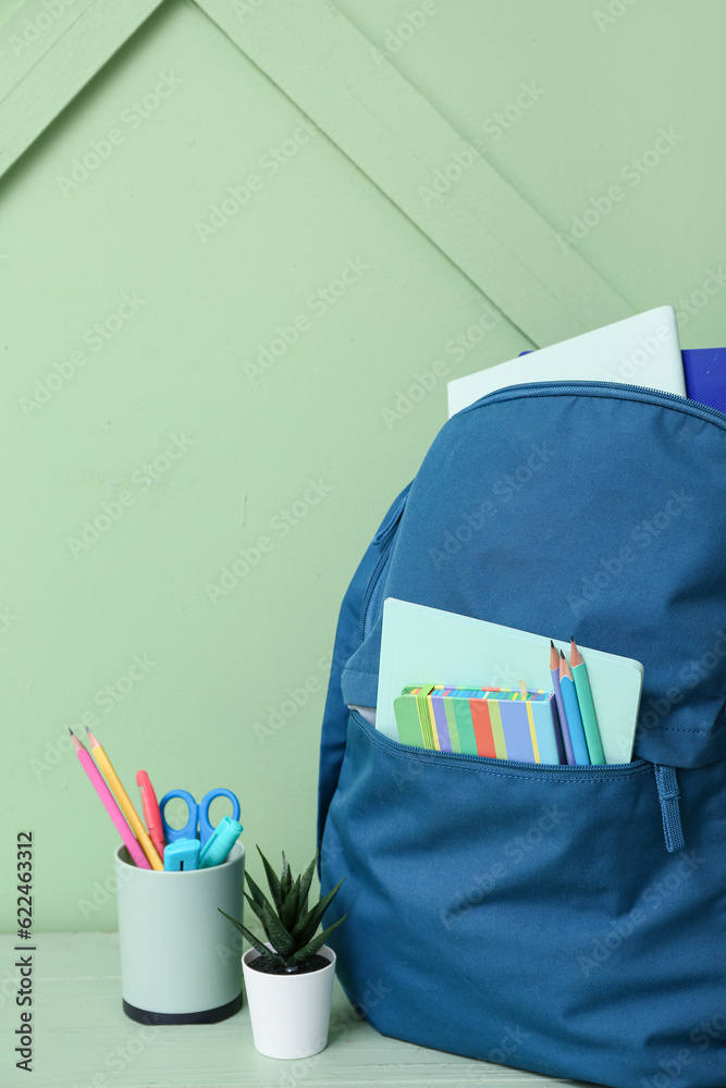 Blue school backpack with notebooks, pencils and houseplant on wooden table near green wall