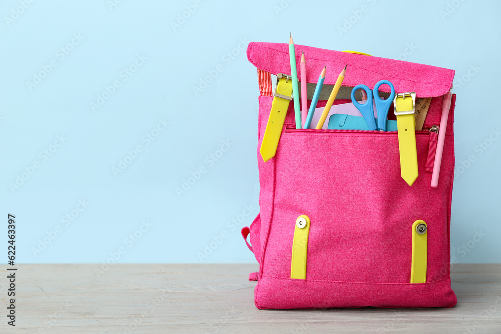 Pink school backpack with scissors, notebooks and pencils on grey wooden table near blue wall