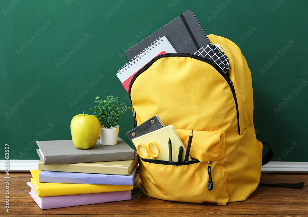 Yellow school backpack with books, houseplant and apple on brown wooden table near chalkboard