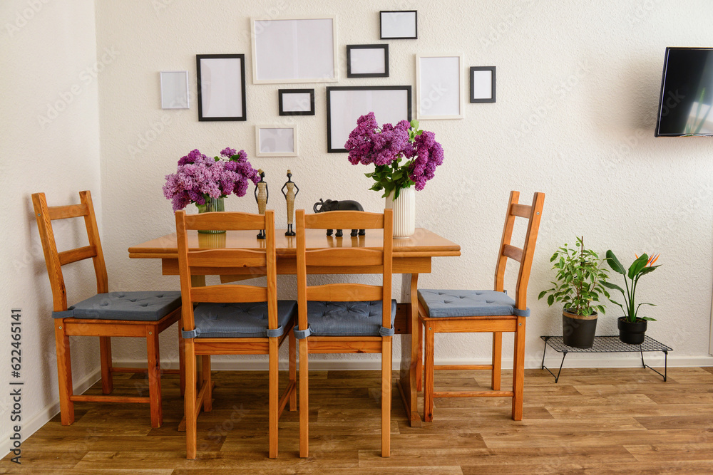 Vases with beautiful lilac flowers on table, chairs and blank pictures in interior of light living r