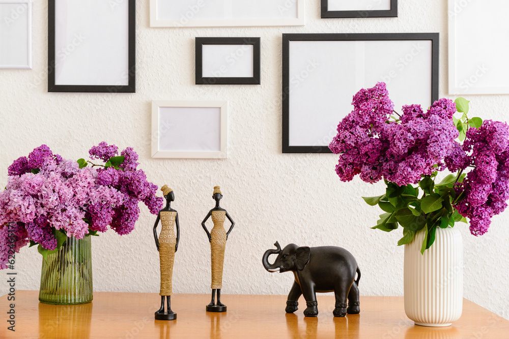 Vases with beautiful lilac flowers, decor on table and blank pictures in interior of light room, clo