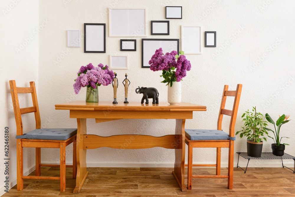 Vases with beautiful lilac flowers on table, chairs and blank pictures in interior of light living r