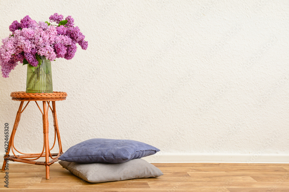 Vase with beautiful lilac flowers on table and pillows near light wall in room