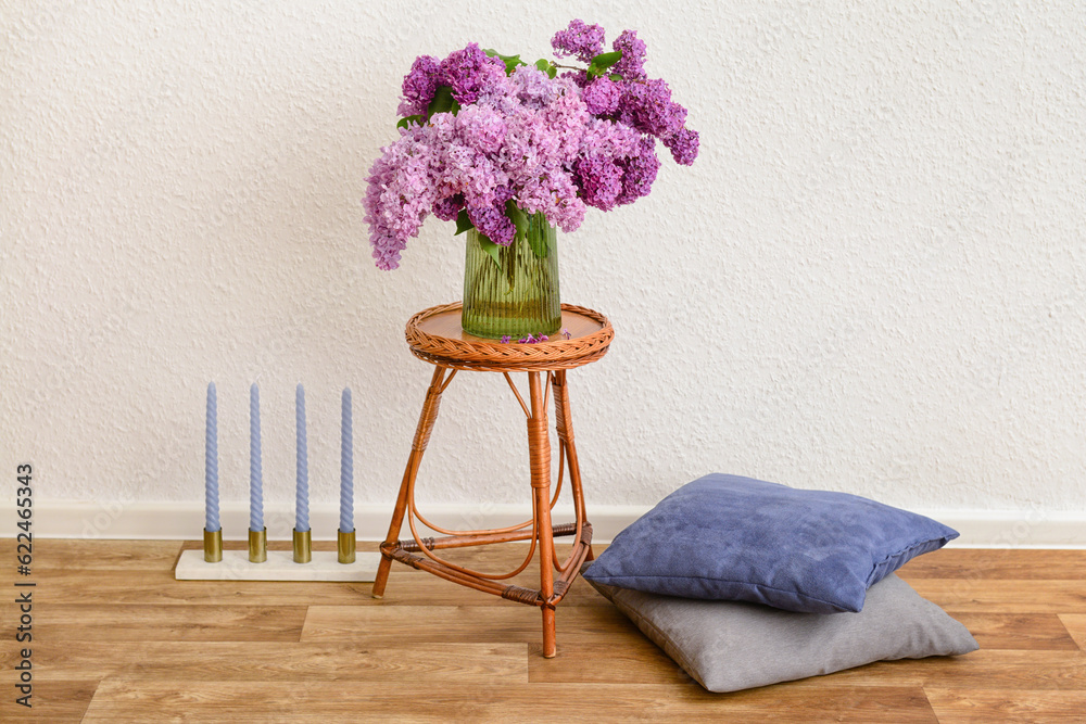 Vase with beautiful lilac flowers on table, pillows and candles near light wall in room