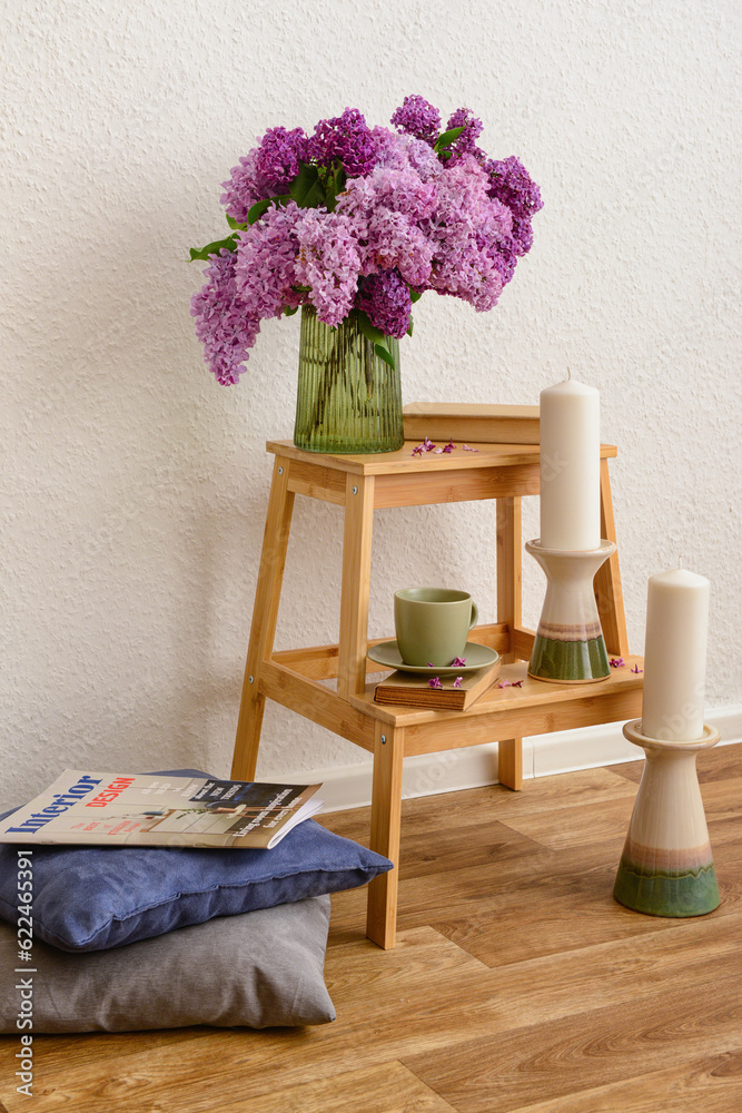 Vase with beautiful lilac flowers, cup on step ladder, pillows and candles near light wall in room