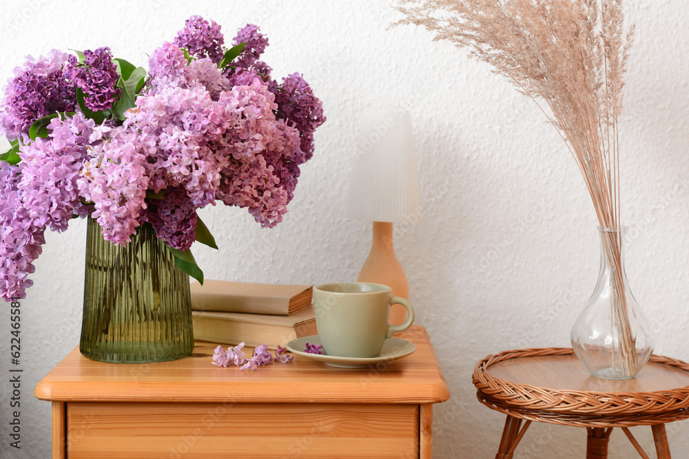 Vase with beautiful lilac flowers, cup on table and pampas grass in room, closeup