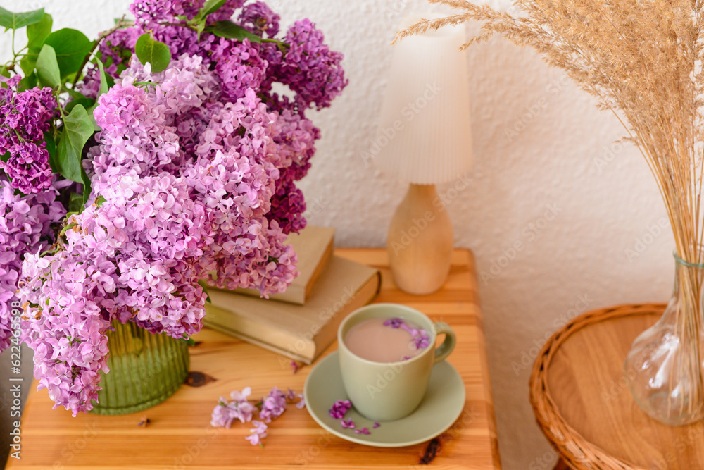 Vase with beautiful lilac flowers, books, cup of coffee on table and pampas grass in room, closeup