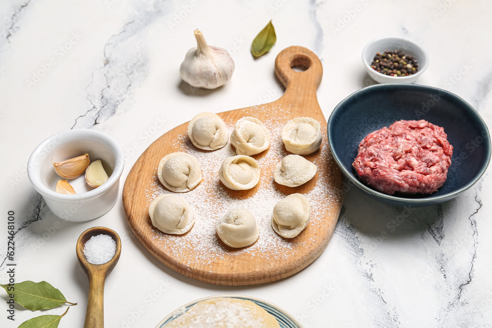 Wooden board with uncooked dumplings and ingredients on white marble background