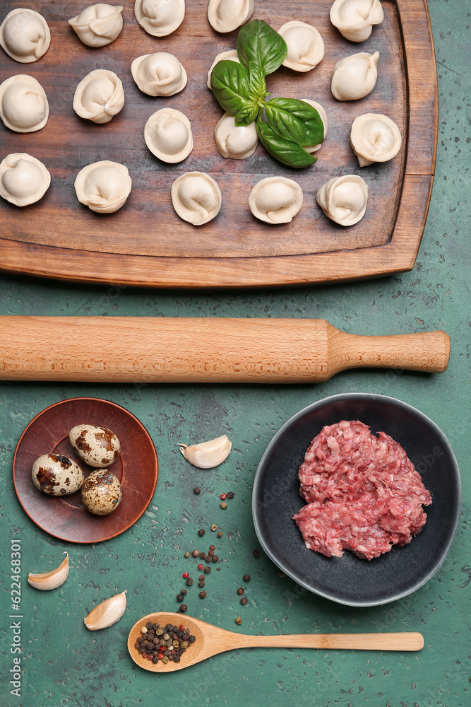 Wooden board with uncooked dumplings and ingredients on green background