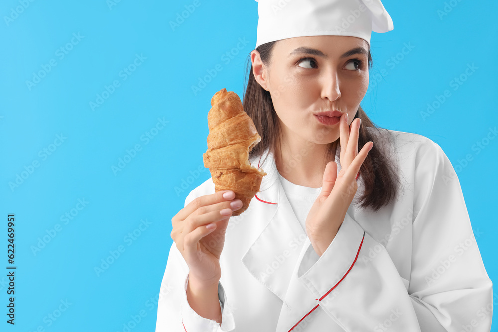 Female chef eating tasty croissant on blue background