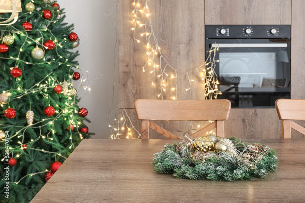 Interior of modern kitchen with Christmas tree, glowing lights and wreath on table