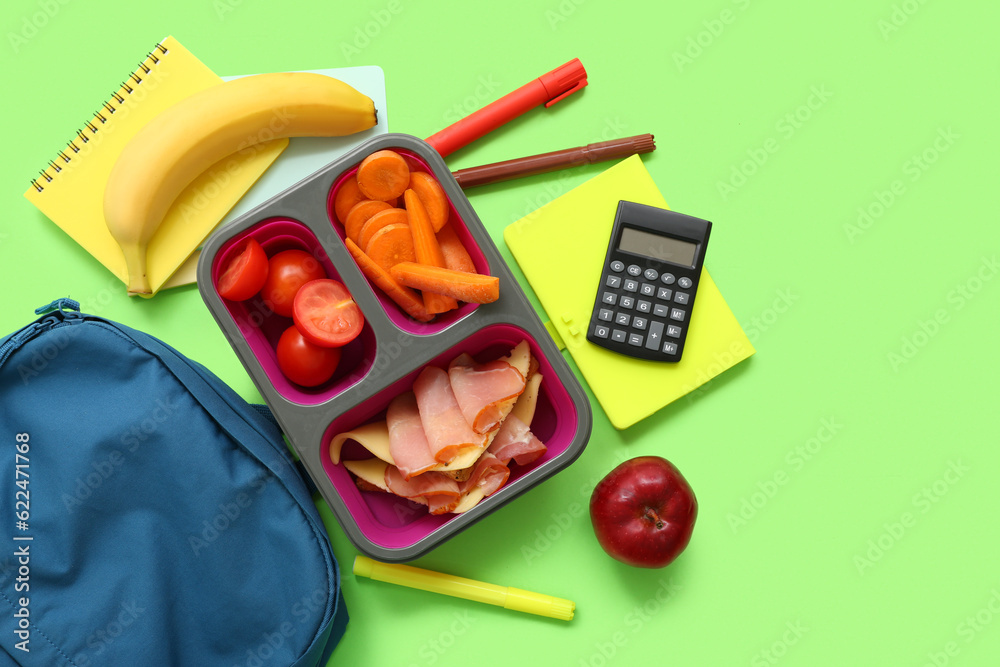 Backpack, stationery and lunch box with tasty food on green background