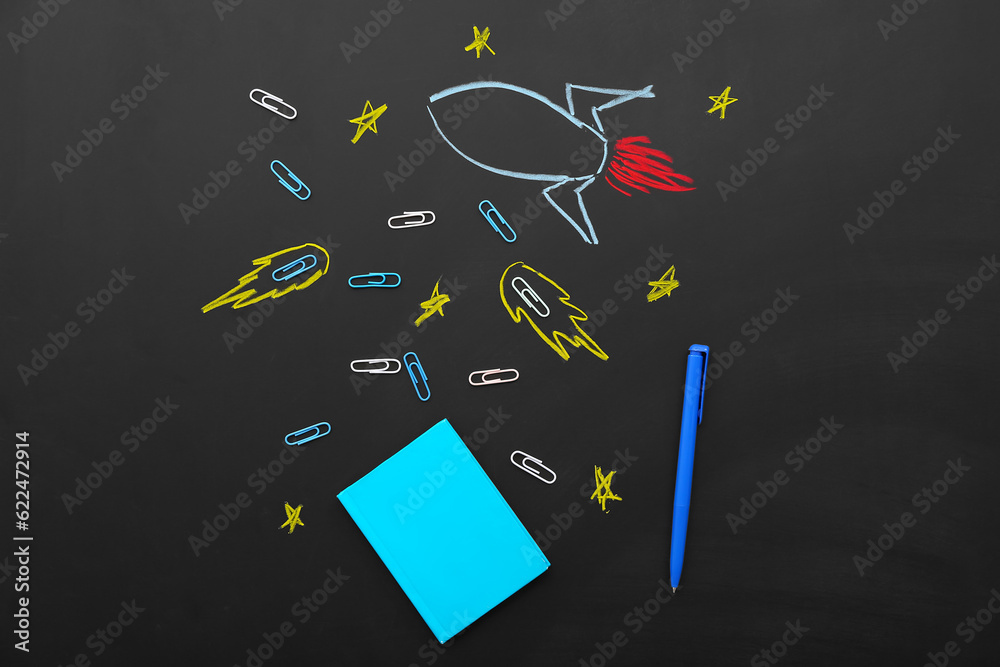 Creative composition with drawn rocket and different stationery on black chalkboard