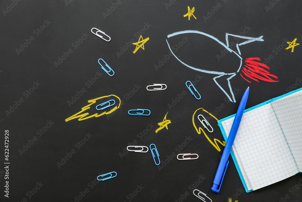 Creative composition with drawn rocket and different stationery on black chalkboard