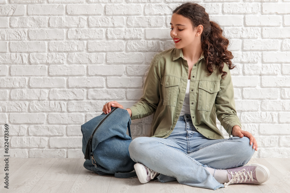 Female student with backpack sitting near white brick wall