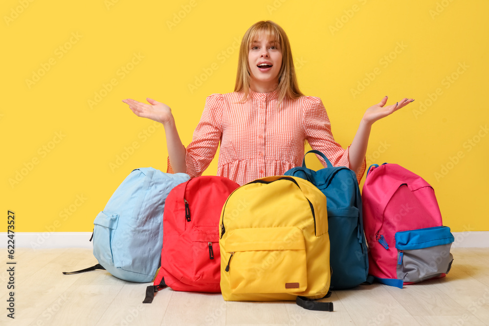 Surprised female student with backpacks sitting near yellow wall