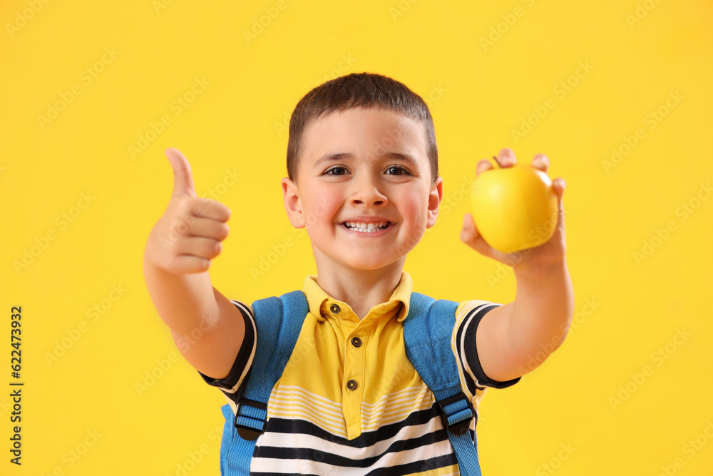 Little schoolboy with apple showing thumb-up on yellow background