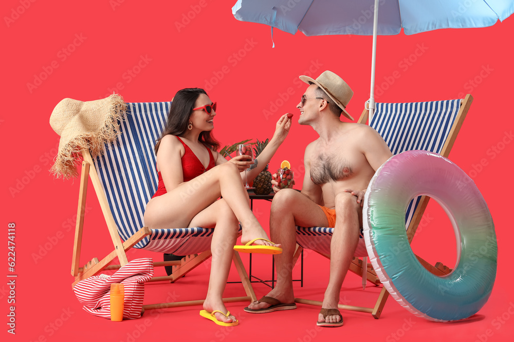 Young couple with strawberries in deck chairs and beach accessories on red background